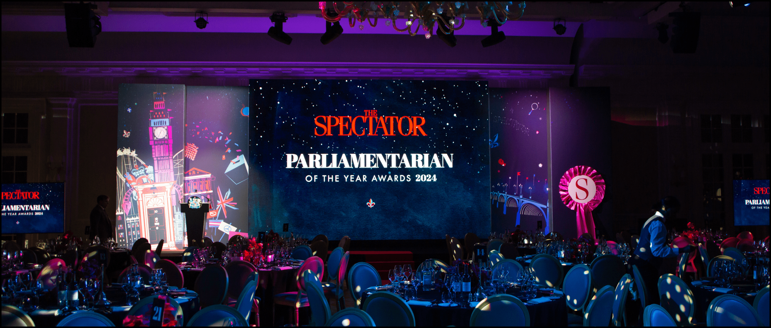 An image displaying London Filmed's Event Production services. A vibrant event venue featuring a fully branded stage with a large LED screen displaying "The Spectator Parliamentarian of the Year Awards 2024." The stage is framed with artistic panels showcasing iconic London imagery, including Big Ben and Westminster.