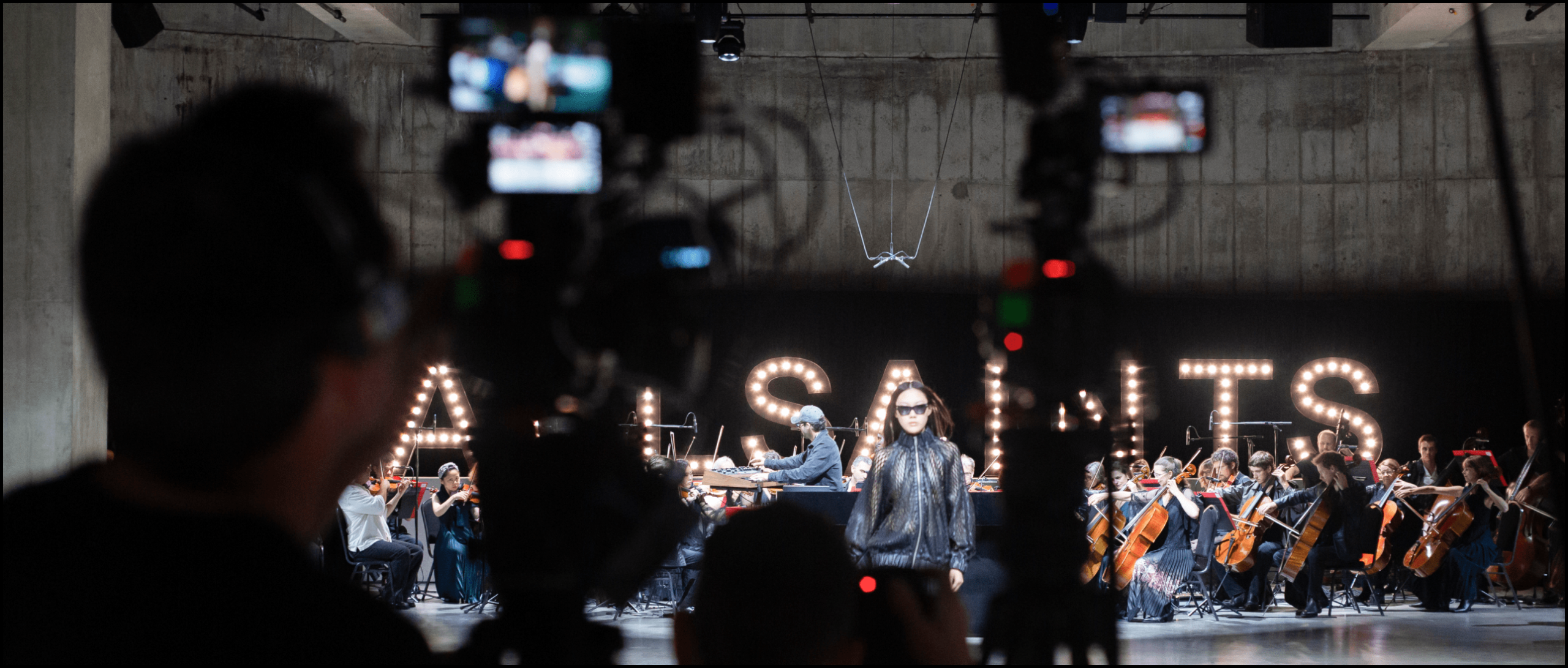 All saints runway 2026 live event showing a live string orchestra with models and the behind the scenes camera set up